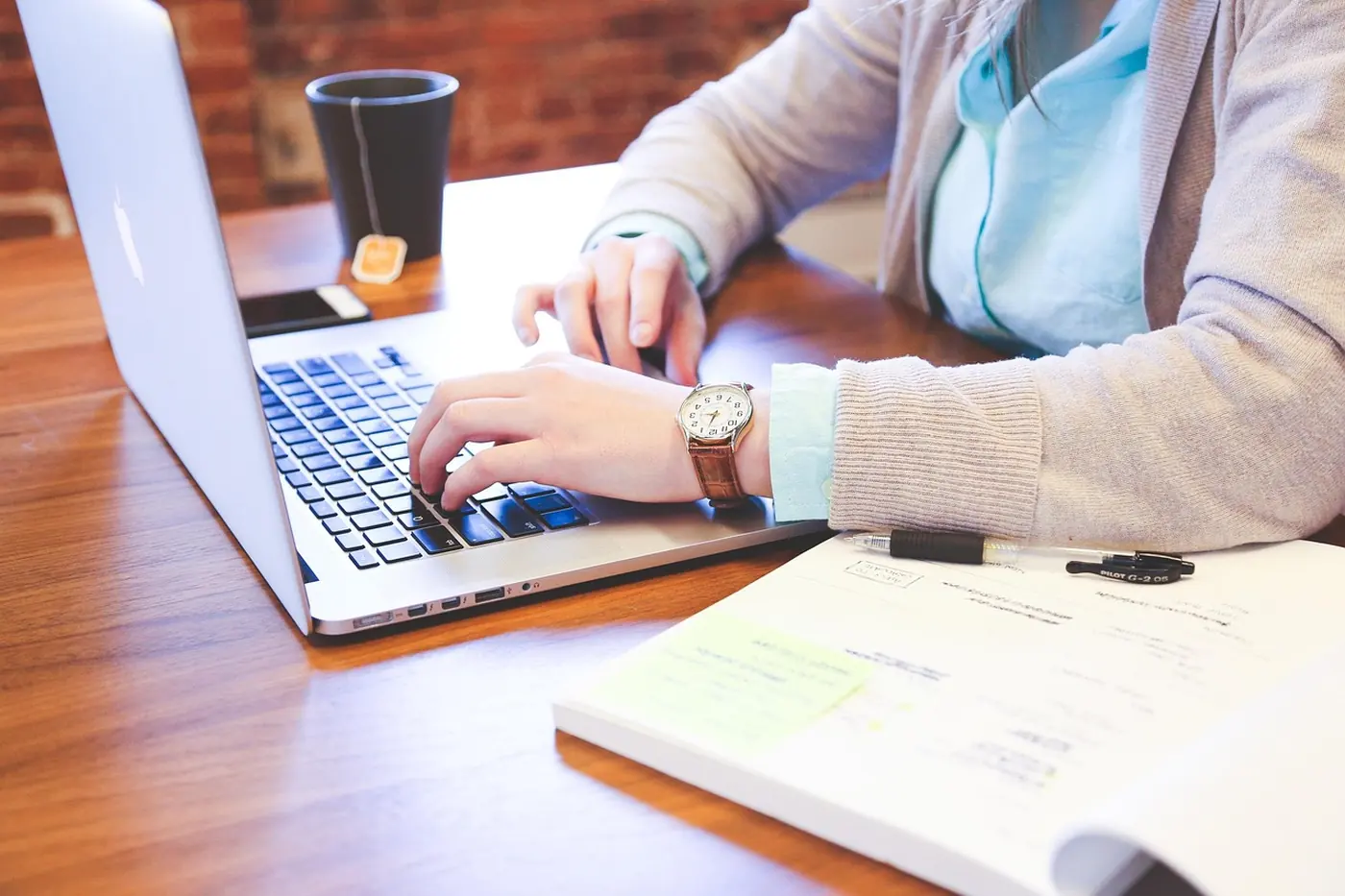 Hands typing on a laptop at a wooden desk beside an open notebook with highlighted notes, a pen and a cup of tea