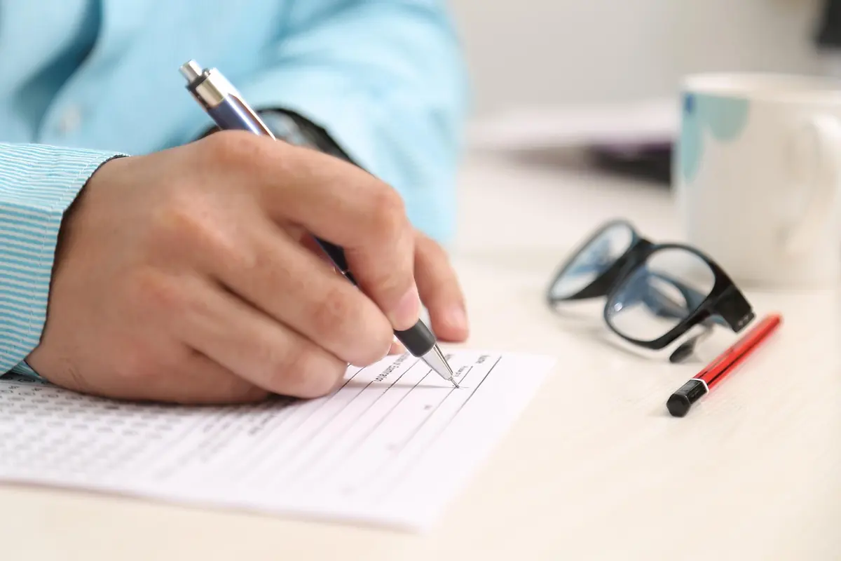 Close-up of a hand ticking lines on a printed form with a pen, with glasses and a red marker lying on the desk