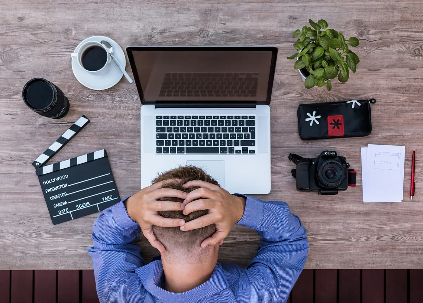 Man seen from above at a wooden desk, holding his head in both hands in front of an open laptop, with a coffee cup, a camera and a film clapperboard around him