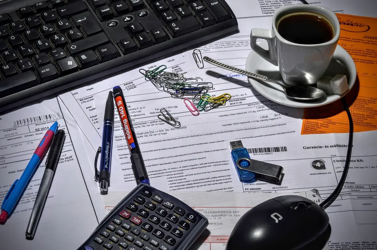 Desk covered with printed invoices, a calculator, pens, paper clips, a USB stick and a cup of coffee next to a keyboard